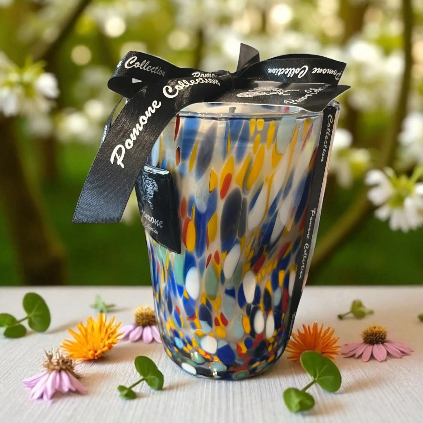 Bougie bleue tacheté orange, rouge et vert, vue de côté avec un arrière plan de fleurs